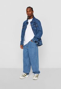 Jeansjacke mit blauen, gerade geschnittenen Jeans, kombiniert mit einem weißen T-Shirt mit rotem Text und weißen Sneakers mit schwarzen und gelben Akzenten.