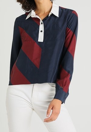 Kvinde iført en marineblå og vinrød langærmet top med hvid krave og knapper, kombineret med hvide bukser, stående mod en ensfarvet baggrund.