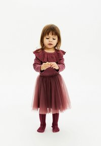 Bordo dolgi rokav s čipkastim ovratnikom z volančki, v kombinaciji s plisejsko krilom iz tila v usklajeni barvi ter rebrastimi bordo geniškimi nogavicami do kolen.
