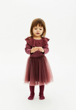 Maglietta bordeaux a maniche lunghe con scollatura di pizzo arricciato, abbinata a una gonna in tulle a strati dello stesso colore e calze al ginocchio a costine bordeaux.