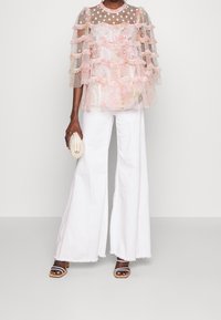 Lyserød, flæset, gennemsigtig bluse med blomsterprint og prikket halsudskæring, parret med hvide, brede bukser og brune sandaler med blokhæle.