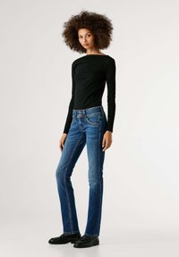 Melnas garām piedurknēm krekls kombinācijā ar zilām izplatītām džinsu biksēm ar pogu akcentiem un izbalējušu tekstūru, valkātas ar melniem apaviem.