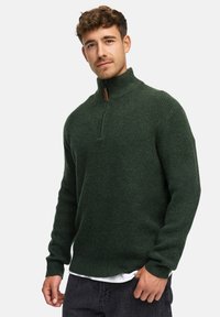 Mann iført en mørkegrønn halv-zip genser og svarte bukser, stående mot en enkel hvit bakgrunn.