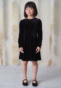 Junges Mädchen mit glatten schwarzen Haaren, das ein langärmeliges schwarzes Samtkleid und schwarze Mary Jane Schuhe trägt, vor einem neutralen Hintergrund stehend.