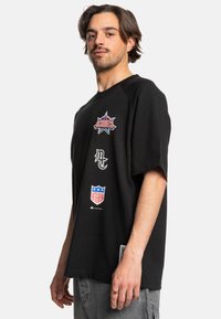 Černé tričko s krátkým ruk�ávem, které obsahuje několik grafických potisků. Uvolněný střih s raglánovými rukávy a jemnou bavlněnou texturou.