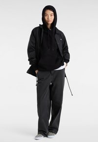 Veste coupe-vent noire sur un sweat à capuche noir, pantalon gris ample et chaussures à enfiler à carreaux. Design simple, tissu doux, coupe décontractée.