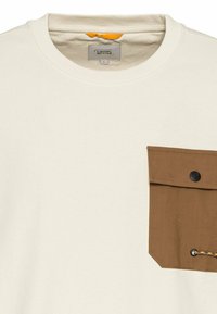 Crème-kleurig T-shirt met een bruine zak die een drukknoop en koorddetail heeft. Zacht materiaal met een geribde halslijn en label binnenin.