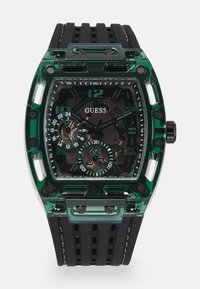 Reloj Guess con caja rectangular de plástico verde, correa de goma texturizada negra y múltiples esferas con números y acentos en negrita.