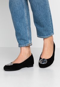 Person med blå jeans og sorte flade sko med store, runde sølvdekorationer på tæerne, stående på hvidt gulv.