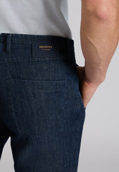 Close-up of person wearing dark blue denim jeans with back pocket and black label reading "INCOTEX BLUE DIVISION."