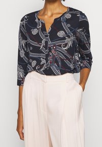 Blusa navy con stampa paisley, caratterizzata da un collo a V e bottoni. Maniche lunghe e tessuto leggero progettato per il comfort. Indossata con pantaloni chiari.