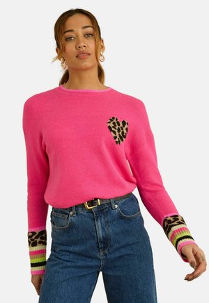 Leuchtend pinker, gestrickter Pullover mit Leoparden-Herz-Motiv, gestreifte Bündchen in Schwarz, Pink und Grün, kombiniert mit hoch taillierter blauer Jeans.