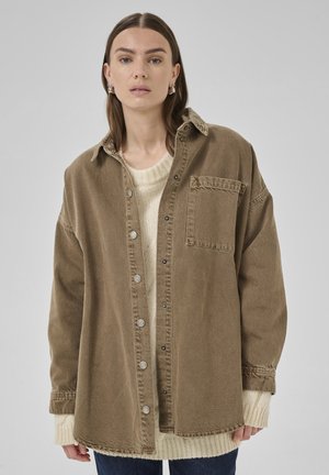 Übergroßes braunes Cordhemd mit großem Kragen, Fronttasche und Druckknöpfen, über einem cremefarbenen Strickpullover getragen. Blaue Jeans vervollständigen das Outfit.