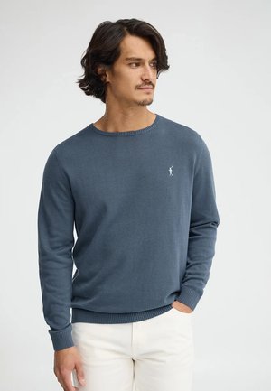 Mand med mellem-langt mørkt hår iført en blå crewneck-trøje med et lille logo og off-white bukser, hånd i lommen, ser til siden.