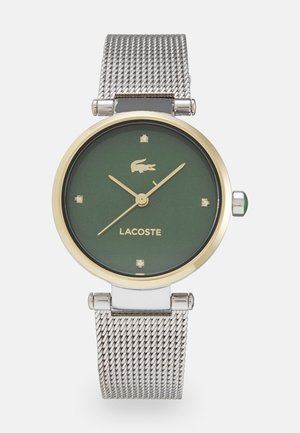 Reloj redondo de Lacoste con esfera verde, bisel dorado, correa de malla plateada, cuatro marcadores de diamante y un logotipo ubicado encima del nombre de la marca.
