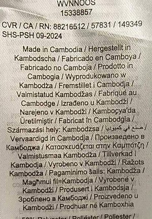 Etichetta con testo multicolore su uno sfondo di tessuto chiaro. Elenca i dettagli di produzione in diverse lingue; indica principalmente "Fatto in Cambogia."