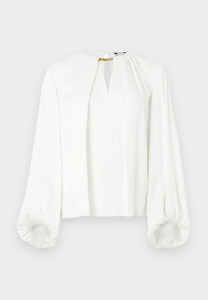 Valkoinen pitkähihainen paita, jossa on avainreikäkaulus, kerätyt olkapäät ja kuminauhalla varustetut hihansuut. Kaulassa kultainen ketju koristeena.