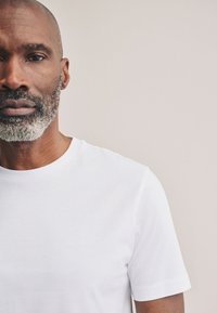 Maglietta bianca in cotone con scollo rotondo, maniche corte, texture liscia e dettagli minimi, indossata da una persona con una barba chiara.