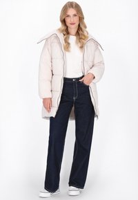 Polstret beige frakke med lynlås, tætsiddende hætte og sidelommer, båret over en hvid skjorte og mørkeblå jeans med kontraststik.