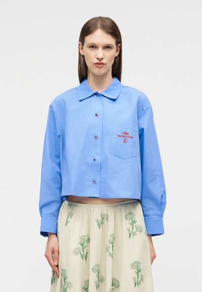 Γυναίκα που φοράει ένα ανοιχτό μπλε κοντό πουκάμισο με κουμπιά, με το λογότυπο "Moje Yacht Club" και άγκυρα στην τσέπη, συνδυασμένο με μια κρεμ φούστα με πράσινη κεντητή λουλουδιαστή διακόσμηση.