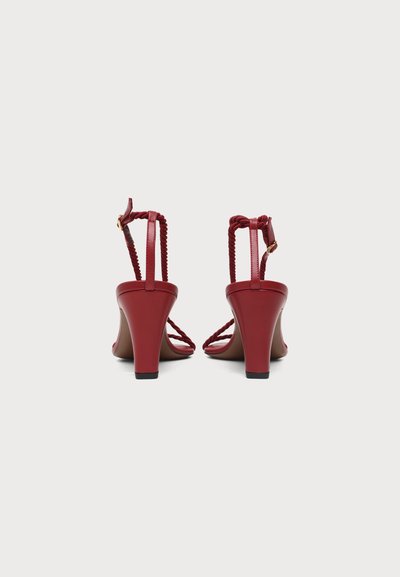 Paire de sandales rouges à talons hauts avec des brides tressées et des boucles à la cheville, vue de dos sur un fond clair uni.