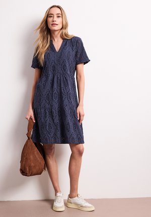 Frau steht an der Wand, trägt ein marineblaues Lochkleid, weiße Sneakers und hält eine braune Ledertasche.