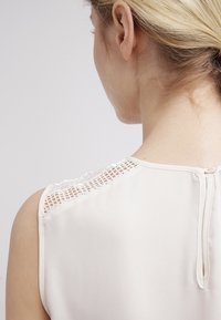 Blouse sans manches beige clair avec un détail en dentelle sur les épaules et une fermeture en forme de clé au dos, fabriquée à partir d'un tissu lisse avec une texture douce.