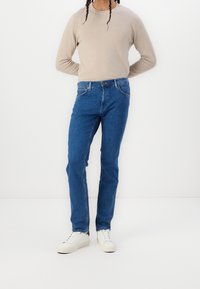 Persona vestida con una camiseta de manga larga beige, jeans azules y zapatillas blancas, de pie contra un fondo blanco liso.