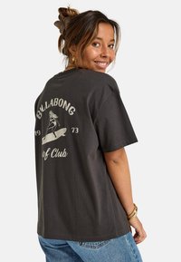 Kvinne som smiler over skulderen iført svart Billabong Surf Club t-skjorte og blå jeans, hår bundet med en stor klype, hvit bakgrunn.