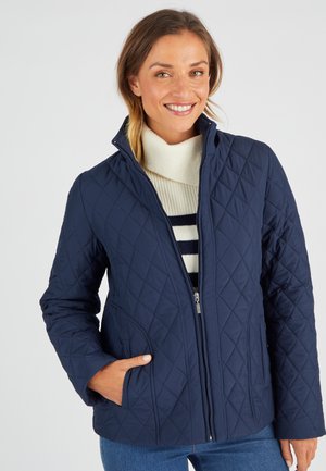 Giacca trapuntata blu navy con colletto alto, chiusura a zip e tasche laterali, abbinata a un dolcevita crema e a uno strato interno a righe blu navy.