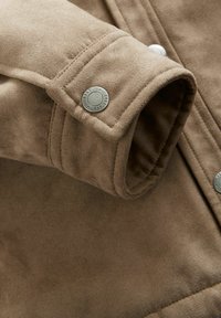 Manșeta jachetei din suede maro cu o cusătură vizibilă și un nasture metalic tip clapetă. Textură moale cu un ușor luciu, având un design pliat.