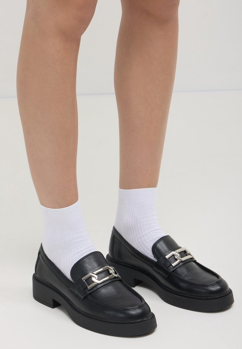 Mocassins en cuir noir avec détails de boucle argentée portés avec des chaussettes blanches côtelées montant à la cheville sur des jambes nues, sur un fond uni.