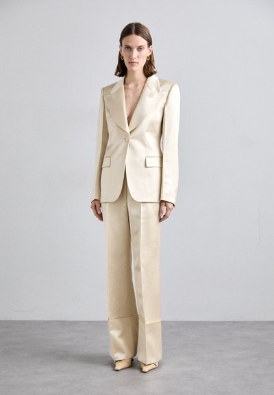 Krämfärgad satinsuit med en djup V-ringad kavaj och vida byxor. Har strukturerade axlar, slag och en minimalistisk design.