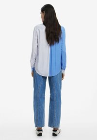 Camicia con colletto e design diviso: una metà a righe verticali blu, l'altra a righe verticali bianche. Vestibilità ampia, maniche lunghe e polsini abbottonati.