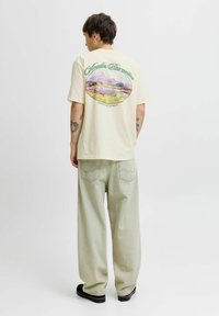 Gaiši beidzēts T-krekls ar krāsainu saulrieta ainavas grafiku uz muguras, savienots ar brīviem zaļganiem džinsiem.