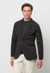 Dunkelgrauer Blazer mit spitzem Revers, einem Knopf und zwei Vordertaschen. Trägt man über einem dunklen Pullover und kombiniert mit beigen Hosen.
