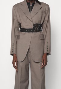 Ljusbrun skräddarsydd kavaj med slag och tre framfickor, åtsnörd med ett svart bälte med silverdetaljer och öglor.