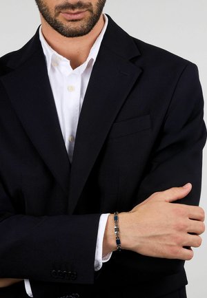 Bracciale in metallo con collegamenti rettangolari blu e argento, indossato su un polso. Il portatore ha indossato una giacca scura sopra una camicia bianca.