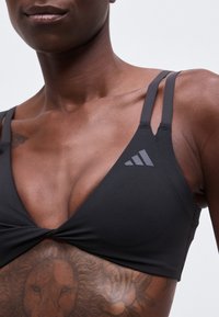 Gros plan du haut du torse d'une personne portant un soutien-gorge de sport noir avec double bretelle et un tatouage visible à l'épaule.