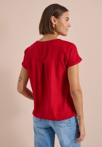 Femme aux cheveux courts bruns, debout, tournée vers la droite, portant un haut rouge à manches courtes et un jean bleu clair sur un fond beige.