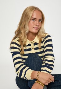 Pruhovaný pletený sveter v žltej a námorníckej modrej, s golierom a tromi gombíkmi. Modelka sa predvádza s džínsami, pričom zdôrazňuje šperky.