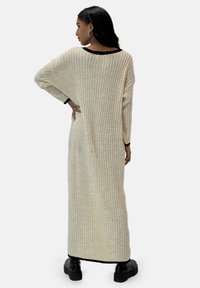 Langes, offenes Strickkleid in Creme mit geripptem Muster, schwarzem Besatz und lockerem Schnitt. Verfügt über eine gerade Silhouette und überschnittene Schultern.