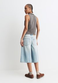 Gecasteter grauer Textur-Ärmelsloser, hellblaue weit geschnittene Jeansshorts und braune Sneakers. Von hinten mit einem schlichten Hintergrund präsentiert.