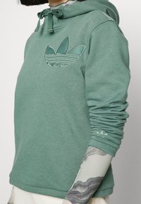 Grön hoodie i mjukt material, med en prägling av en trebladig logotyp på bröstet, ribbade ärmslut och dragsko i huvan.