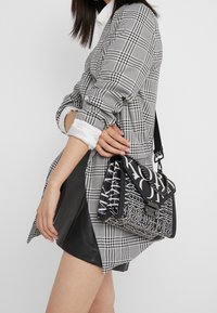 Donna che indossa un blazer a scacchi e una gonna nera, tenerando una borsa a tracolla in bianco e nero con logo Michael Kors.