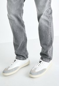 Adidași albi cu accente din suede gri, închidere cu sireturi și o talpă texturată, asortați cu blugi gri din denim într-o frumusețe lejeră.