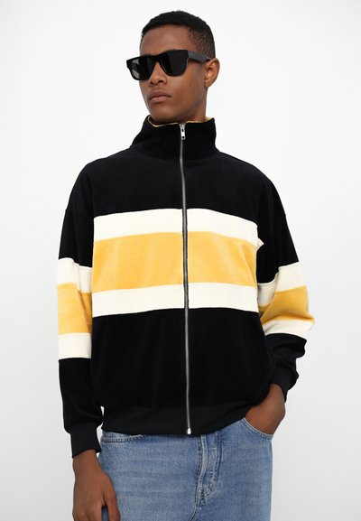 Giacca a zip in velour nero con spesse strisce orizzontali gialle e bianche, collo alto e polsini a coste. Indossata con occhiali da sole neri.