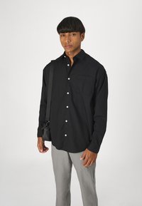 Svart långärmad skjorta med knappar och en framficka, kombinerad med ljusgrå byxor. Modellen bär en liten svart väska över axeln.