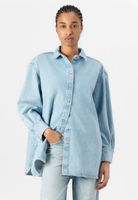 Lyseblå oversized denimskjorte med knaplukning foran, spiss krage og lange ermer, med en subtil slitt tekstur.