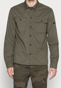 Uomo che indossa una camicia verde oliva con bottoni e due tasche sul petto, con pantaloni camouflage, in piedi con le braccia rilassate lungo i fianchi.